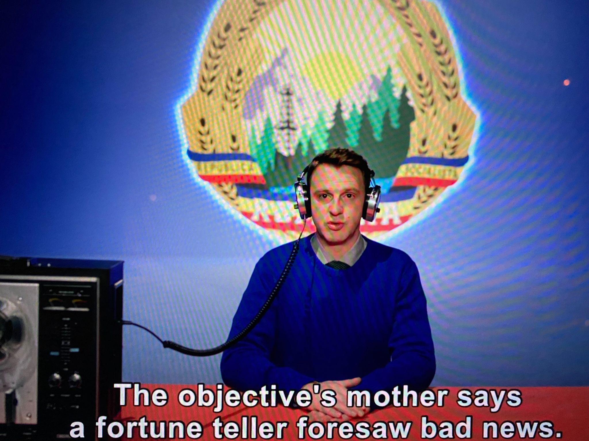 Movie still of a man sitting behind a desk with a radio system. The caption reads "The objective's mother says a fortune teller foresaw bad news."