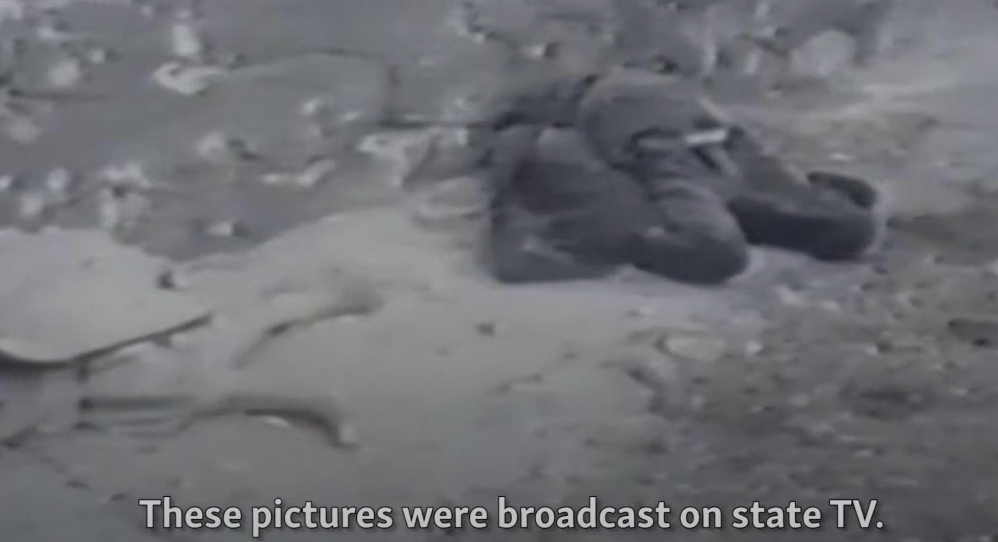 Black and white news image of the dictator and his wife dead in the street after their executions. Bullet holes can be seen on the wall behind them.