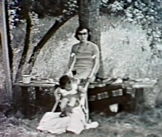 Black and white image of the the author outside as a child sitting in a chair with her mother behind her.