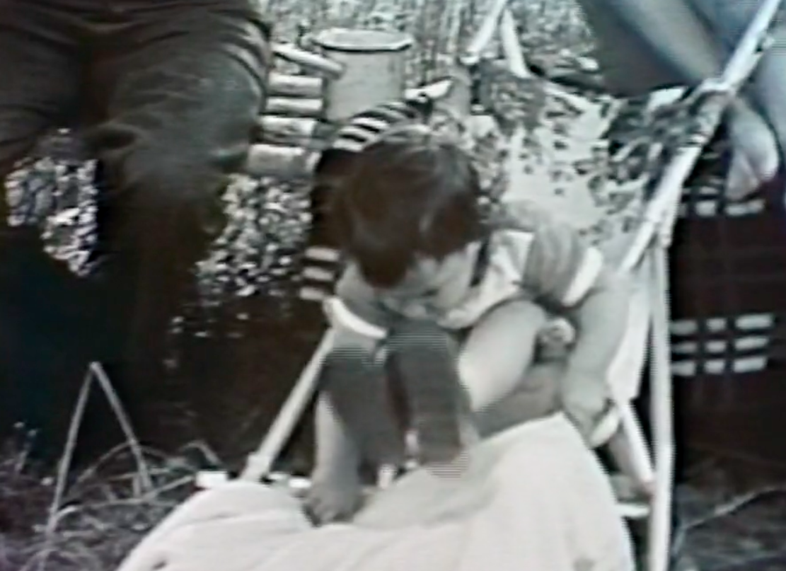 Black and white image of the the author outside as a child sitting in a chair and reaching down to grab a blanket.