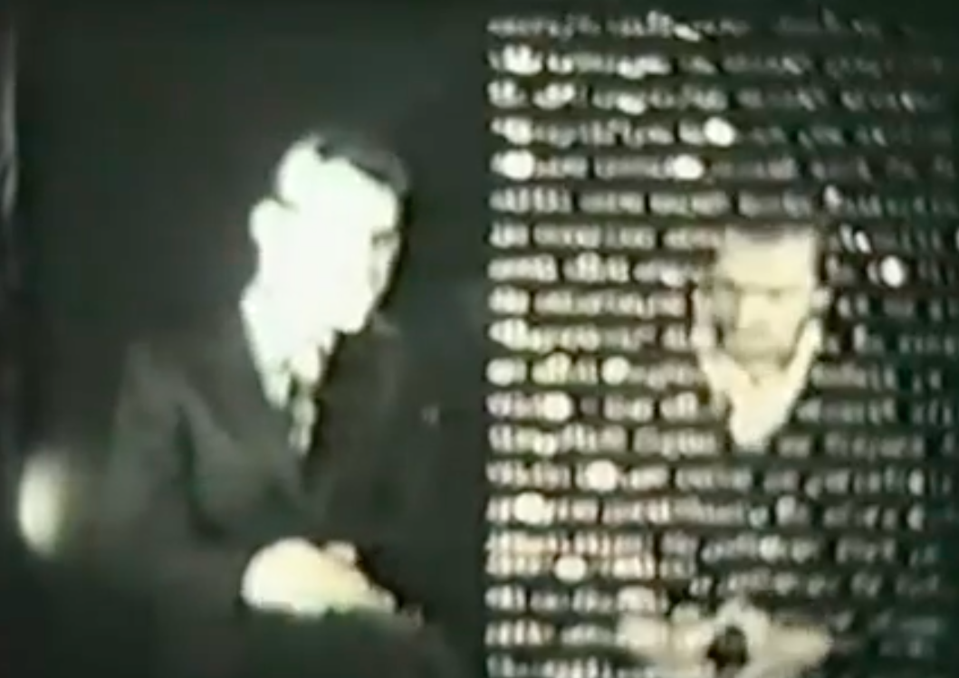 A blurry film still in which two men sit side by side. The man on the right is obstructed by illegible text.